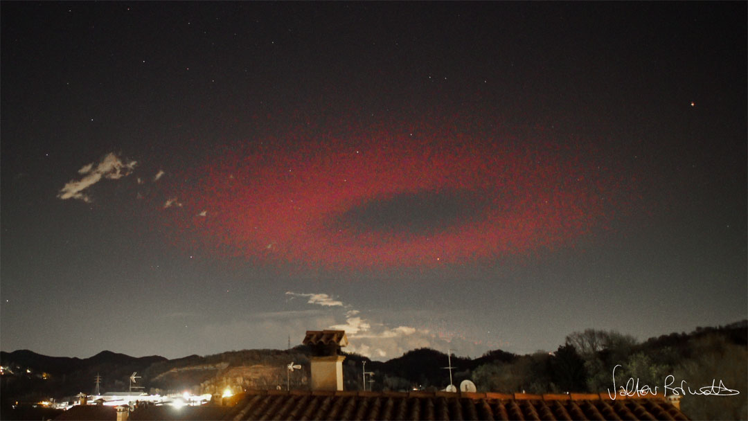 A large red ring is seen high above a landscape that has 
sparse clouds and a foreground building.
Please see the explanation for more detailed information.