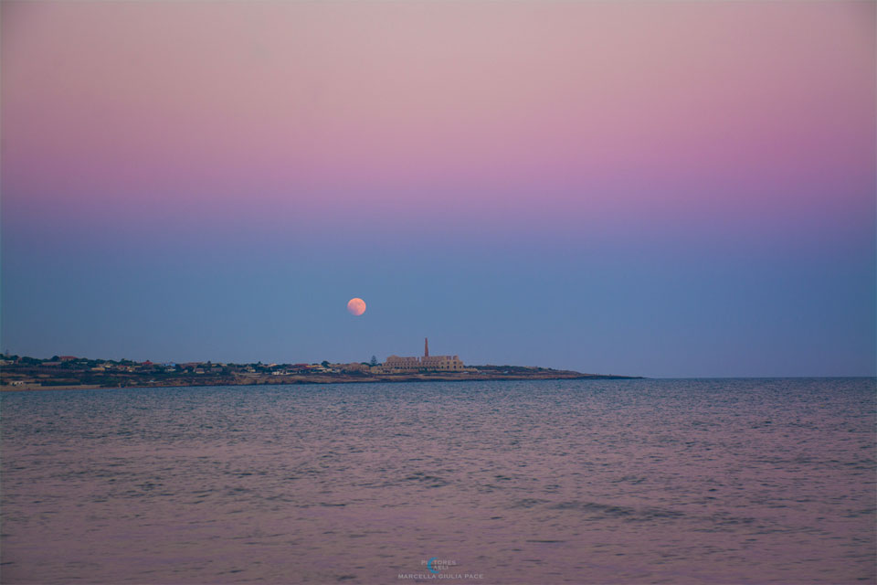 A sunset sky over Sicily is shown with, from top to bottom, a pink 
atmospheric band, a blue atmospheric band containing the Moon, a band
with land containing buildings, and a band of water reflecting the 
pink and blue atmospheric bands.
Please see the explanation for more detailed information.