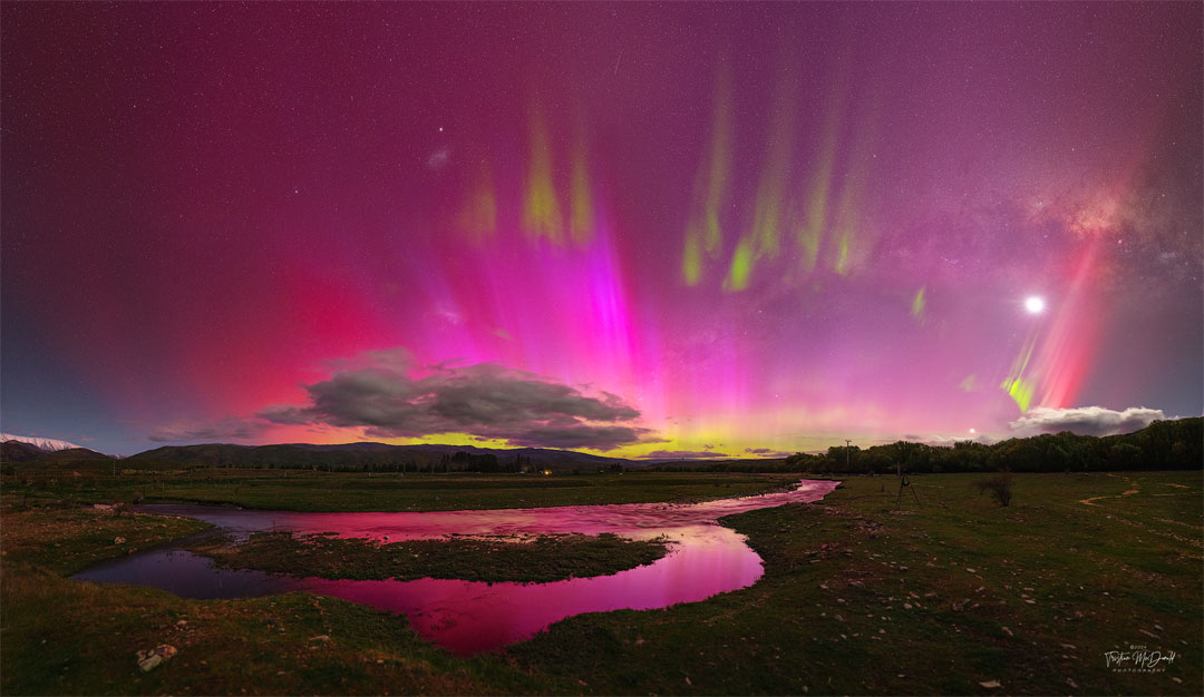 A night sky is shown that appears mostly red due to pervasive
aurora. In the foreground is covered by watery grasslands. Clouds 
are visible above the horizon. Thin green aurora are visible 
toward the top of the frame. In the background one can find the Moon,
the LMC, SMC, Venus, a meteor, and the band of our Milky Way galaxy.
Please see the explanation for more detailed information.