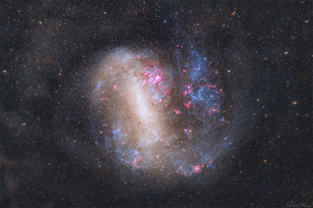 An unusual looking galaxy is shown with a light bar
running nearly vertical and blue stars and red nebulas 
around the edges.  
Please see the explanation for more detailed information.