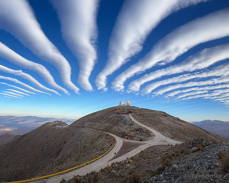 A series of white parallel clouds are seen going off
into the distance in a background blue sky. In the foreground
is a hill with two domes at the top. 
Please see the explanation for more detailed information.